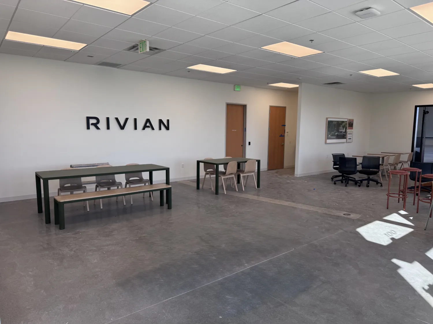 commercial-1 Spacious office with tables, chairs, and “RIVIAN” on the wall, featuring bright lighting and minimal decor.