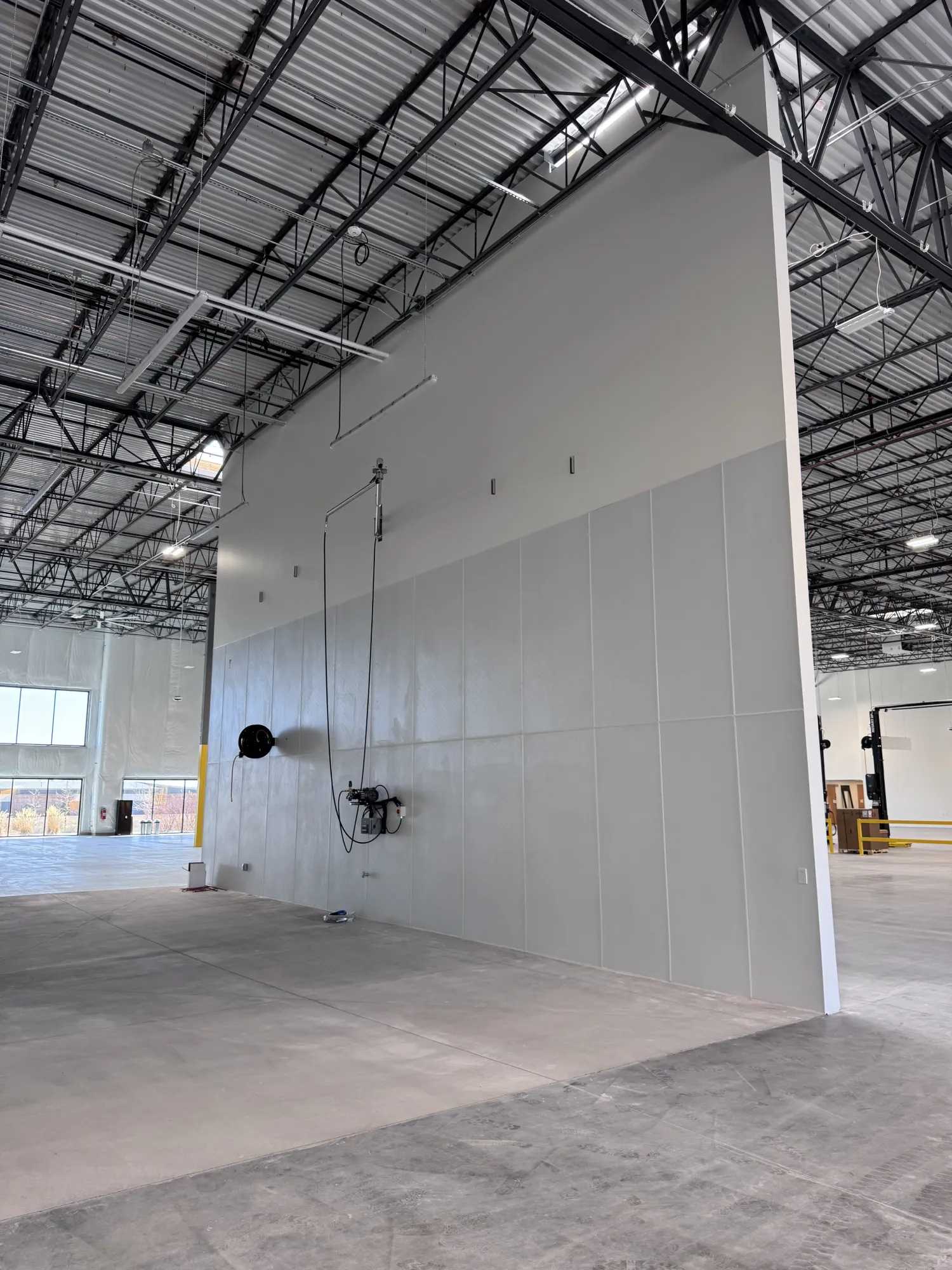 Large, empty warehouse with high ceilings, exposed beams, and a partition wall with hanging cables.