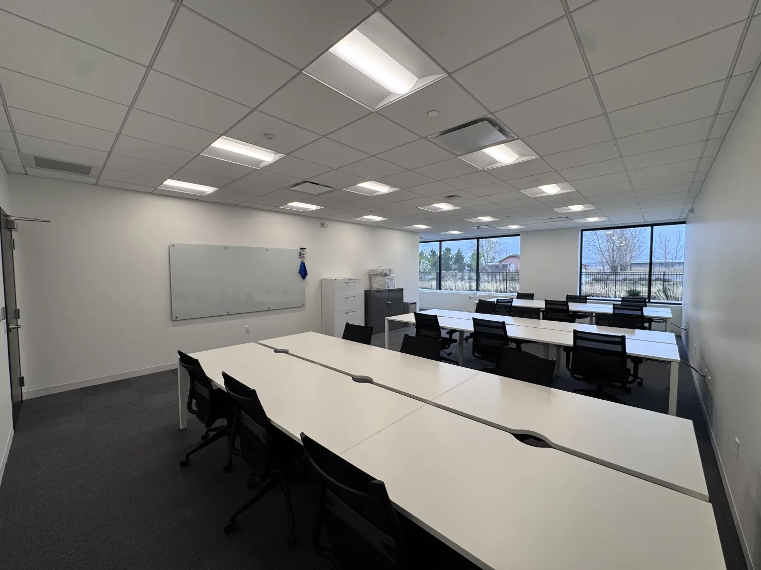 commercial-16 Modern office with white desks, black chairs, large windows, and a whiteboard on the wall.