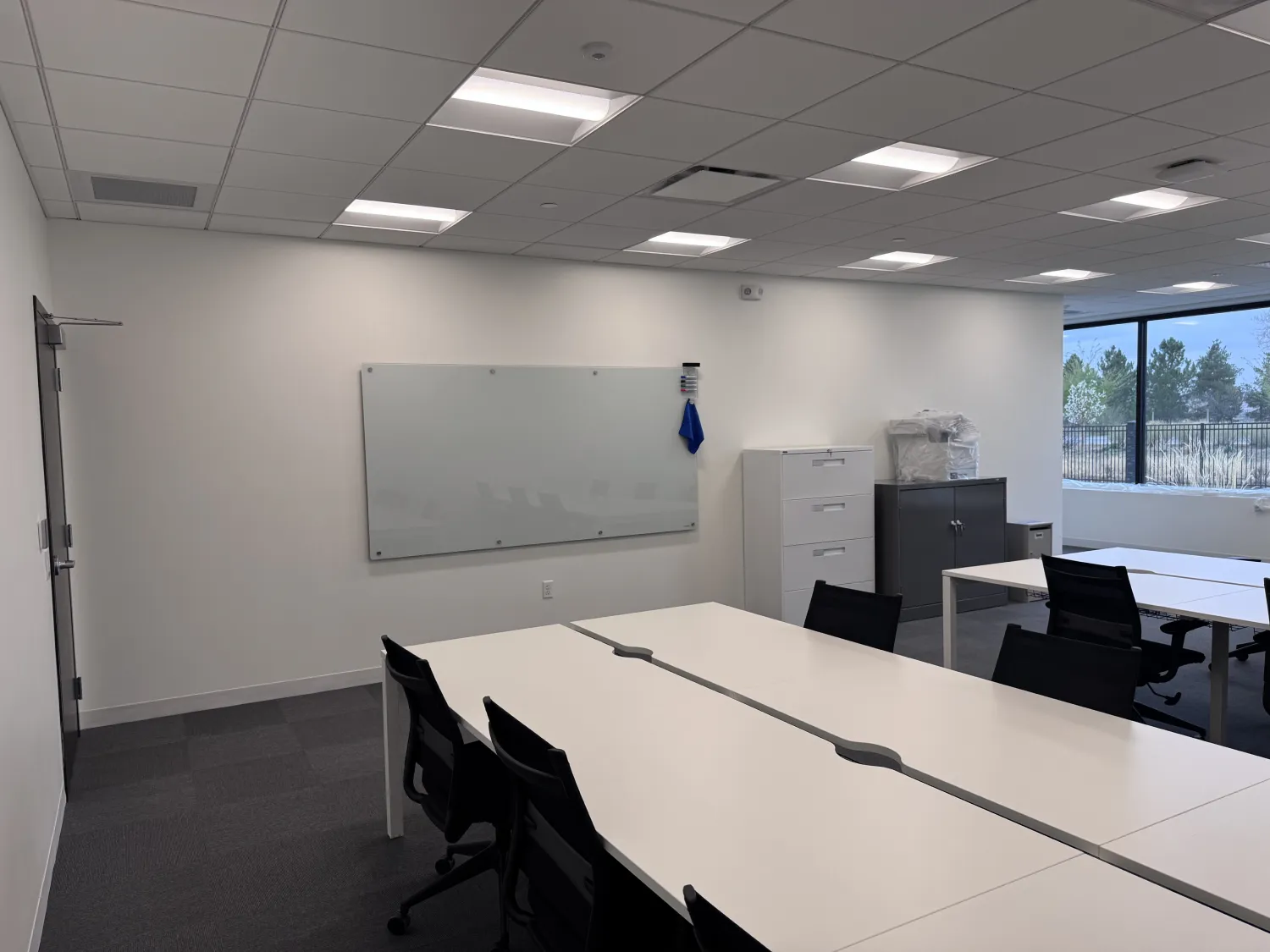 commercial-17 Modern office room with desks, chairs, a whiteboard on the wall, and large windows letting in natural light.