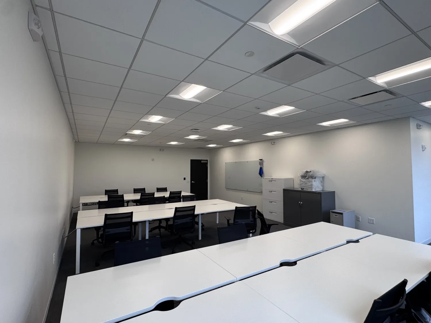 commercial-18 A modern, empty office room with white desks, black chairs, a whiteboard, and cabinets under bright lights.