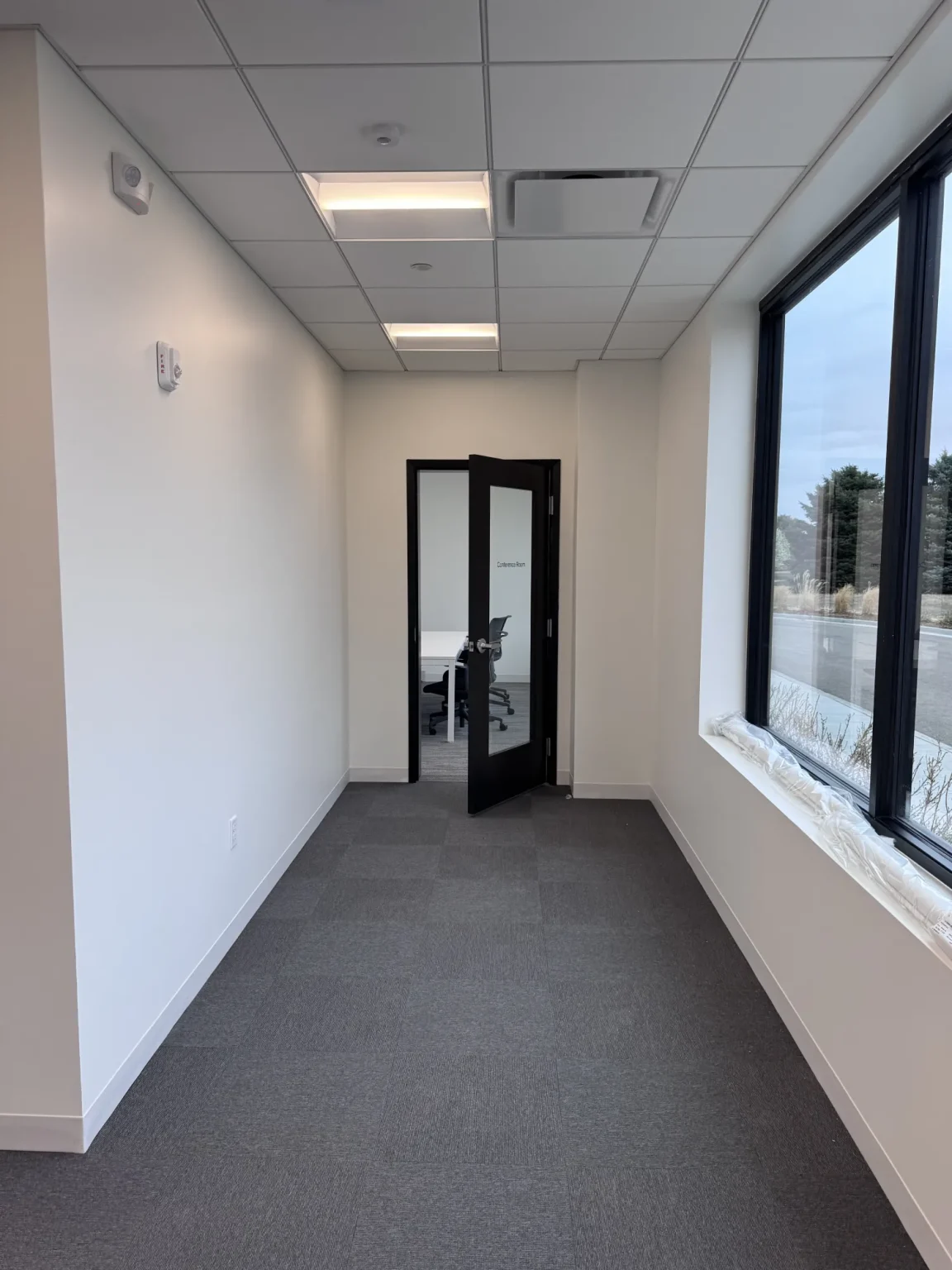commercial-19 A narrow office hallway with gray carpet, white walls, and a glass door leading to a meeting room.