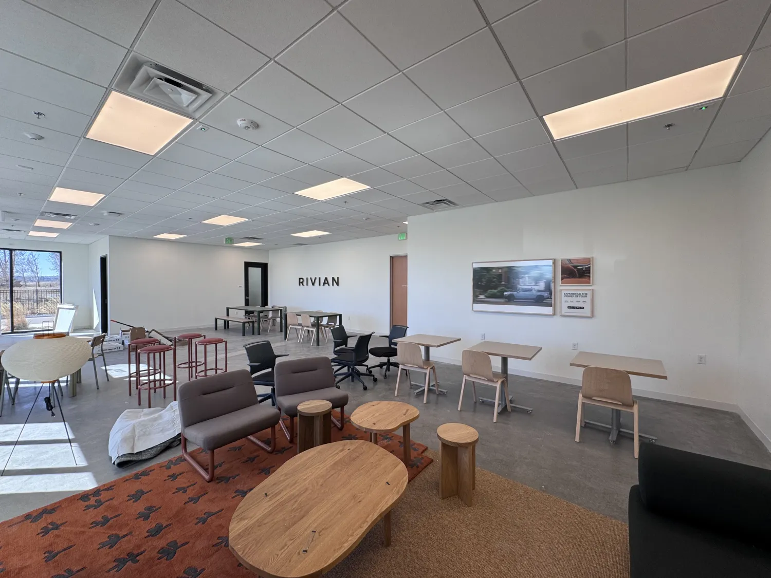 commercial-2 Modern office lounge with tables, chairs, Rivian logo on the wall, and large windows letting in sunlight.