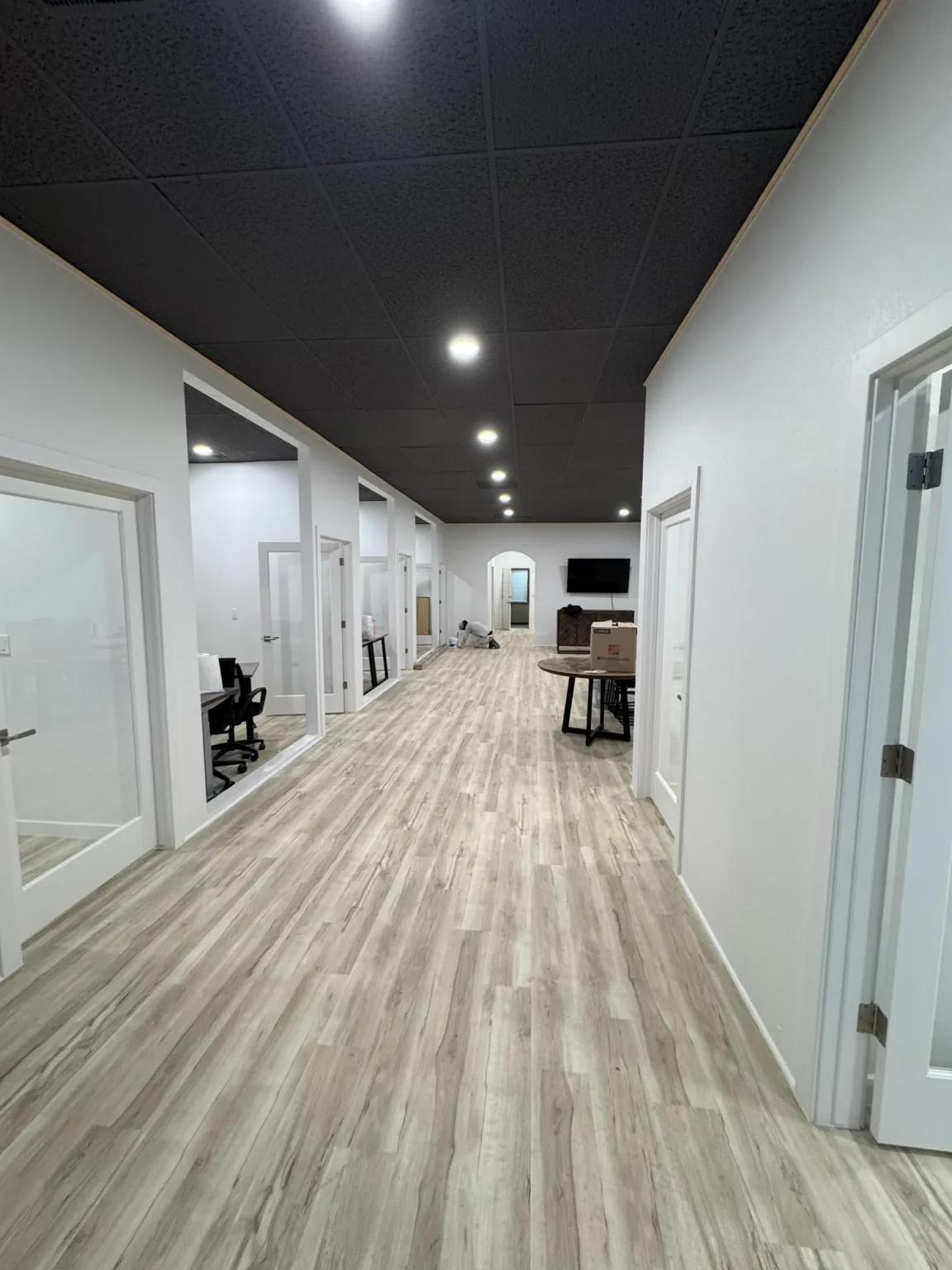 commercial-21 Modern office hallway with wood floor, white walls, glass doors, and desks in separate rooms on the left side.