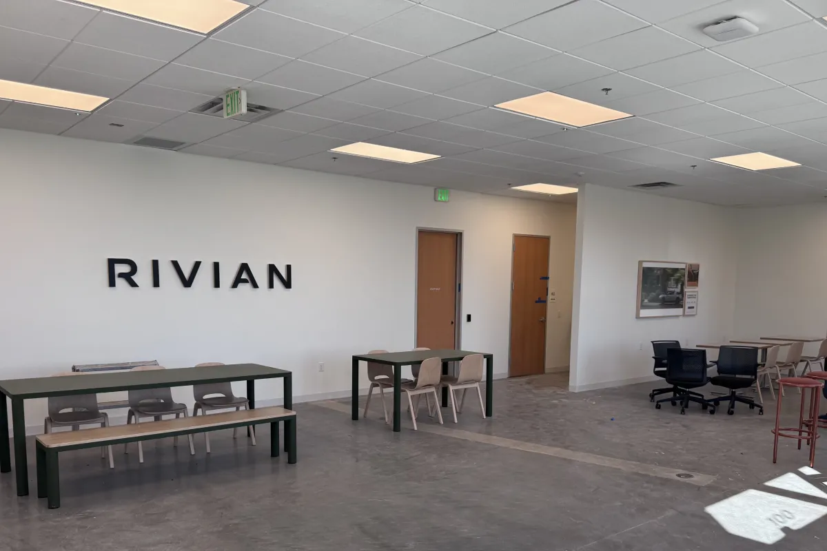 Modern room with "RIVIAN" on the wall, tables, chairs, concrete floor, and two closed wooden doors.
