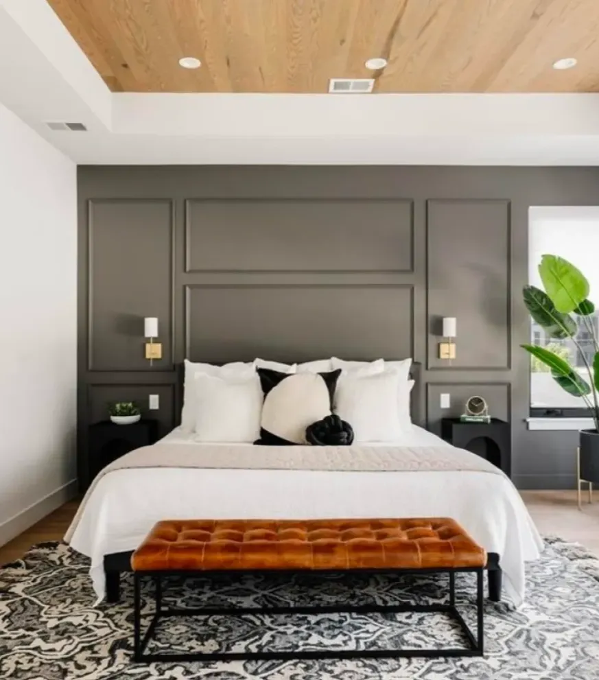 Modern bedroom with a dark accent wall, white bedding, orange bench, plant, and patterned rug.