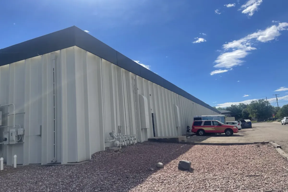 A white industrial building with cars parked outside on a sunny day with a blue sky and some clouds.