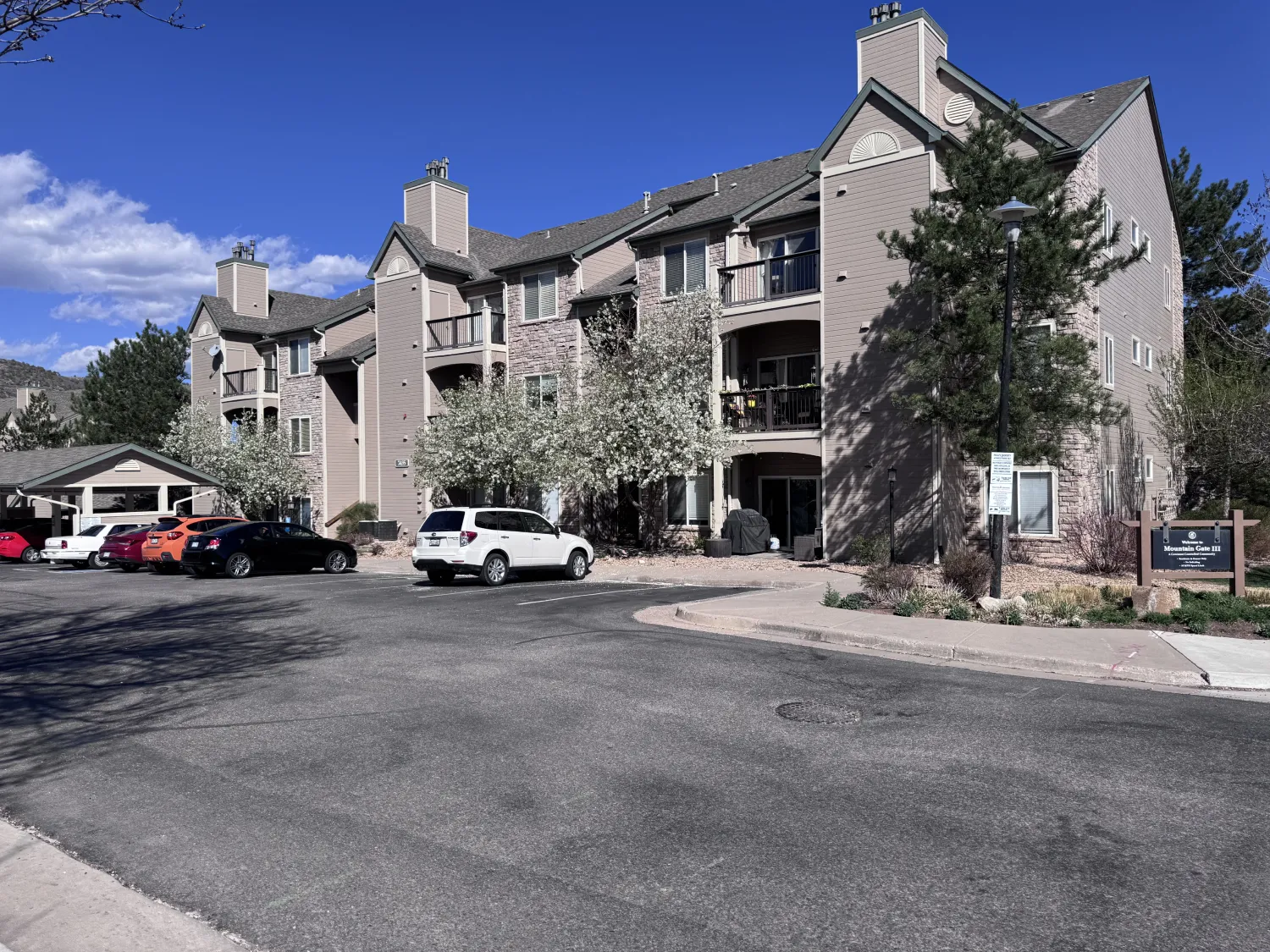 hoa-14 Apartment complex with parked cars, trees in bloom, and a sunny blue sky in the background.