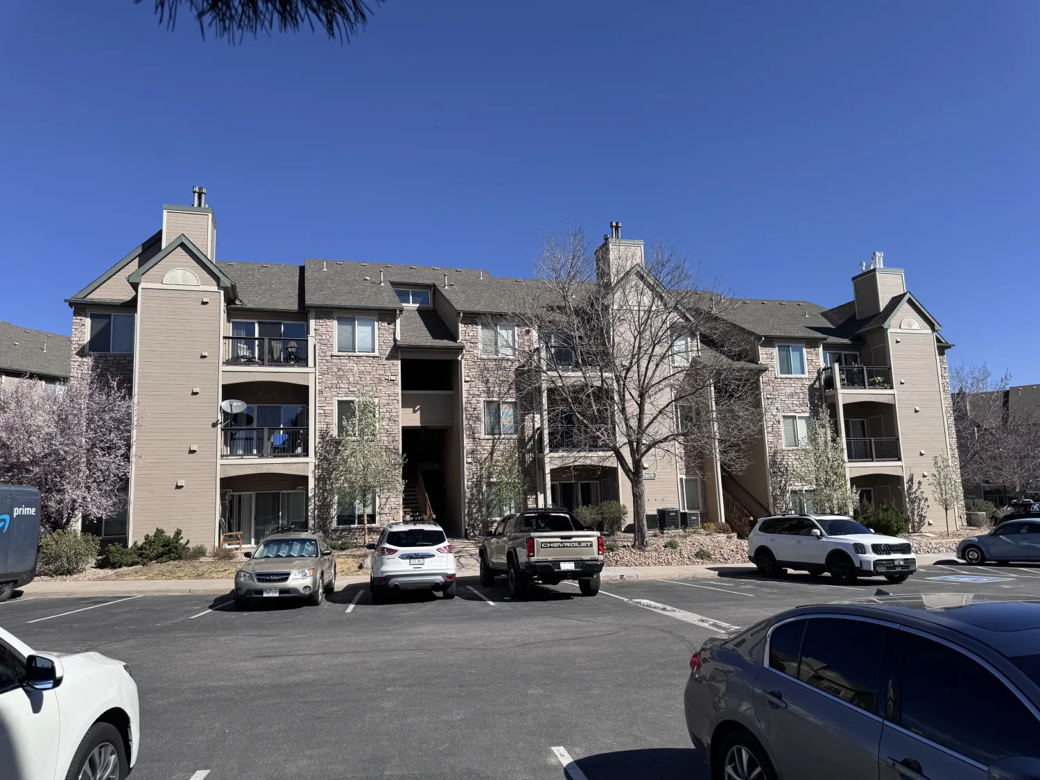 hoa-20 Three-story apartment building with balconies, parked cars, and clear blue sky in the background.