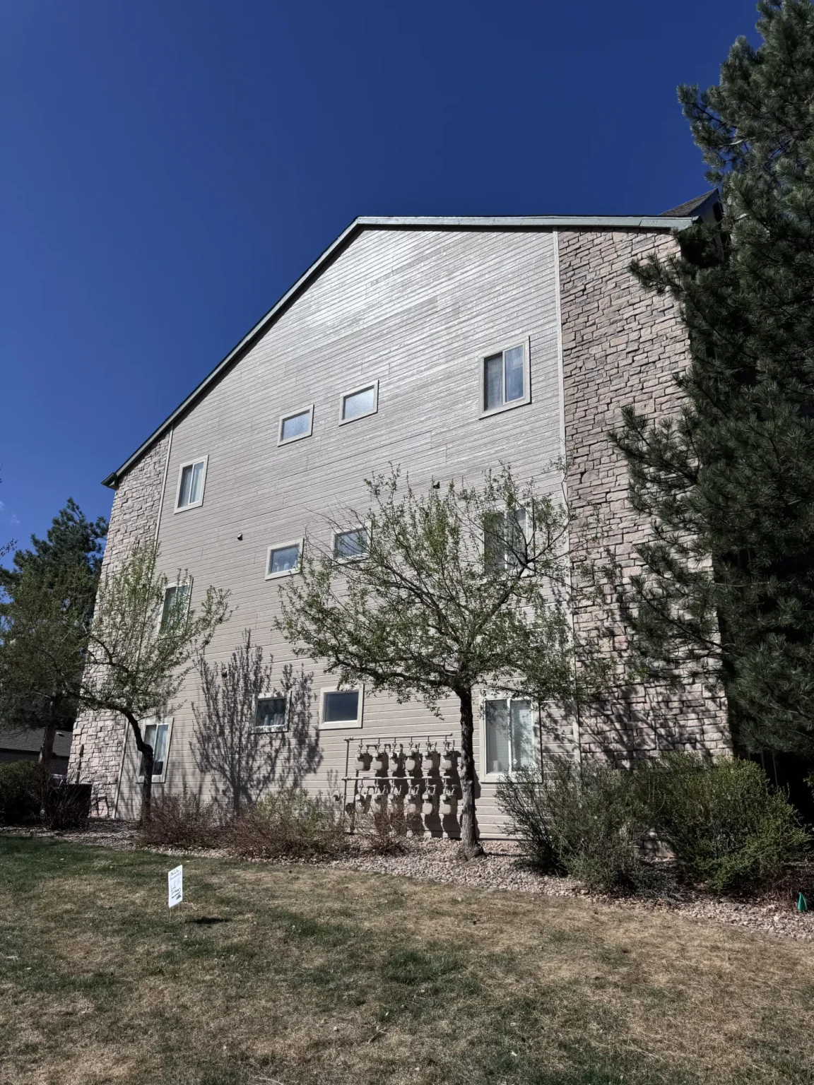 hoa-22 Three-story beige apartment building with stone accents, trees, and grass under a clear blue sky.