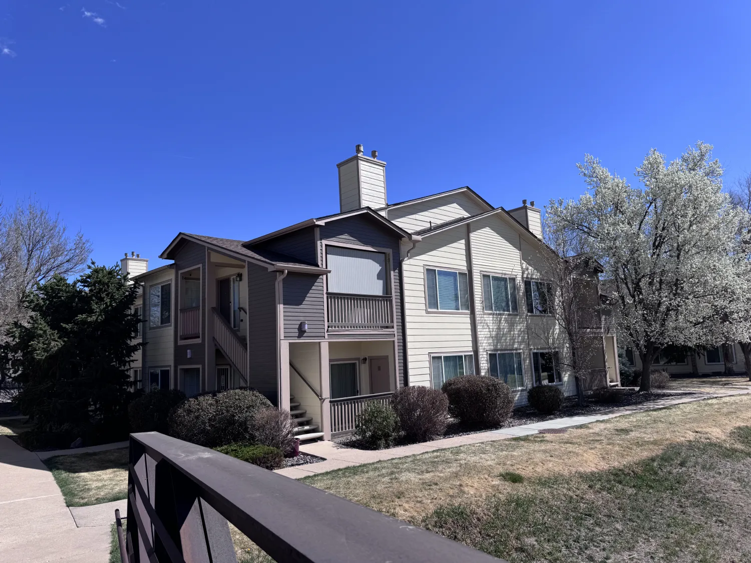 hoa-24 Two-story beige apartment building with balconies, trees, and a grassy area under a clear blue sky.