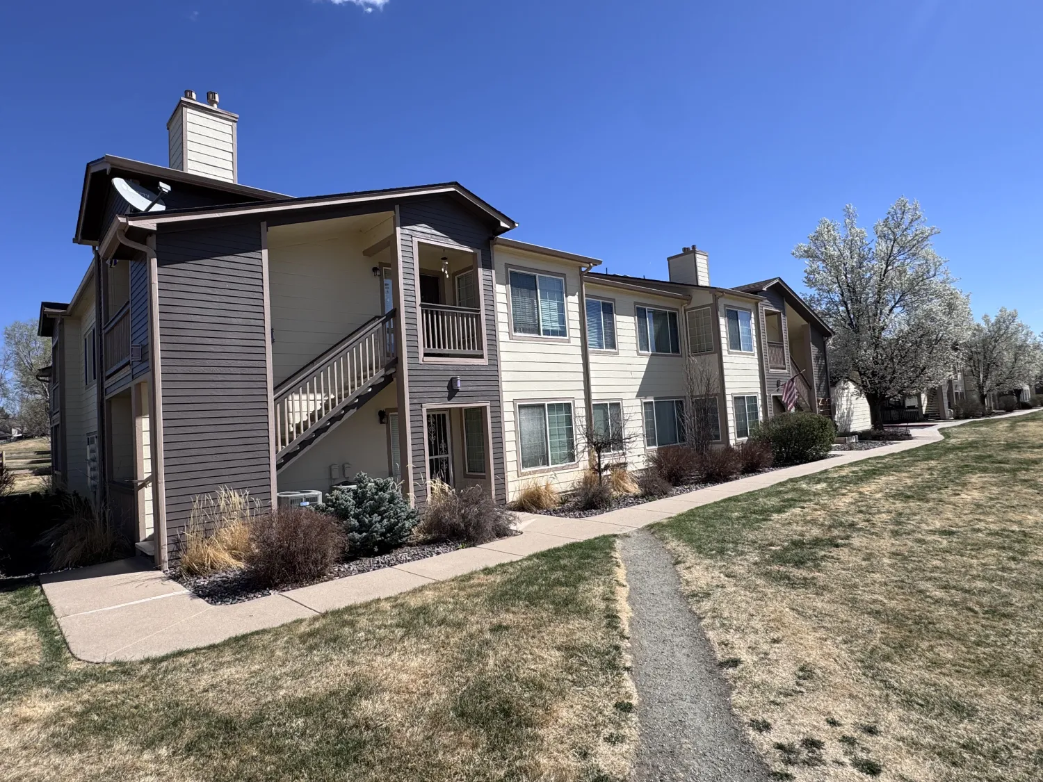 hoa-25 Two-story apartment building with beige siding, outdoor stairs, and a grassy yard under a clear blue sky.