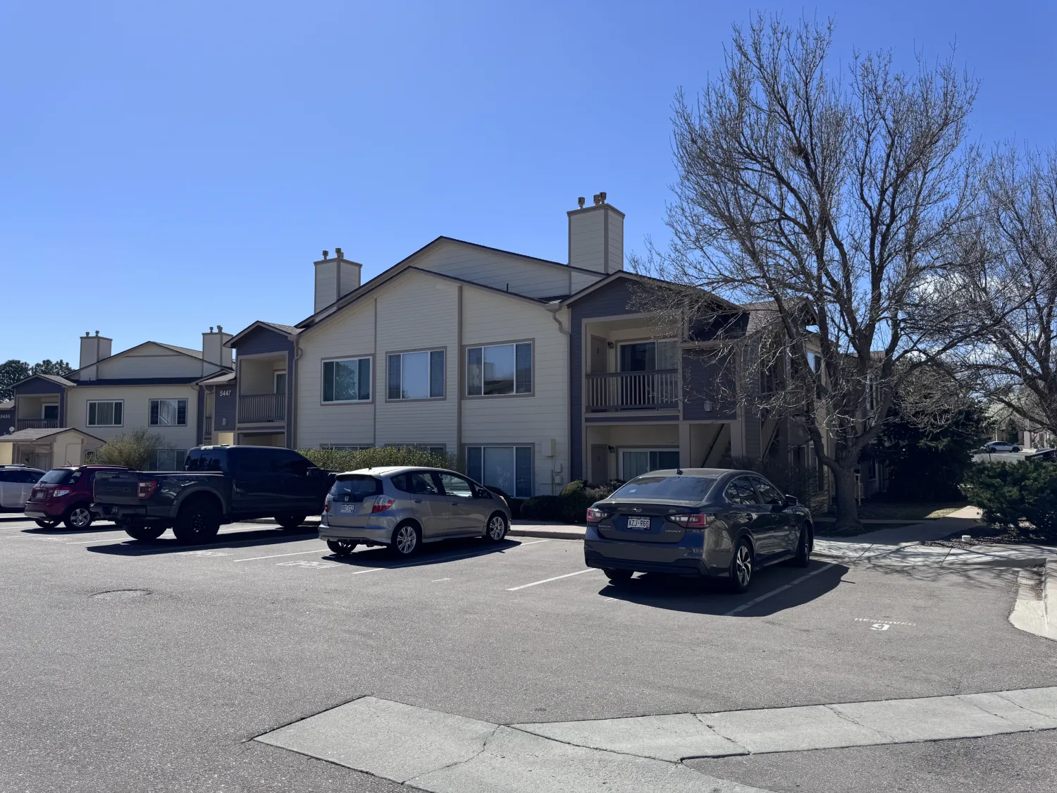 hoa-26 Apartment building with parked cars in front, leafless tree, and clear blue sky on a sunny day.