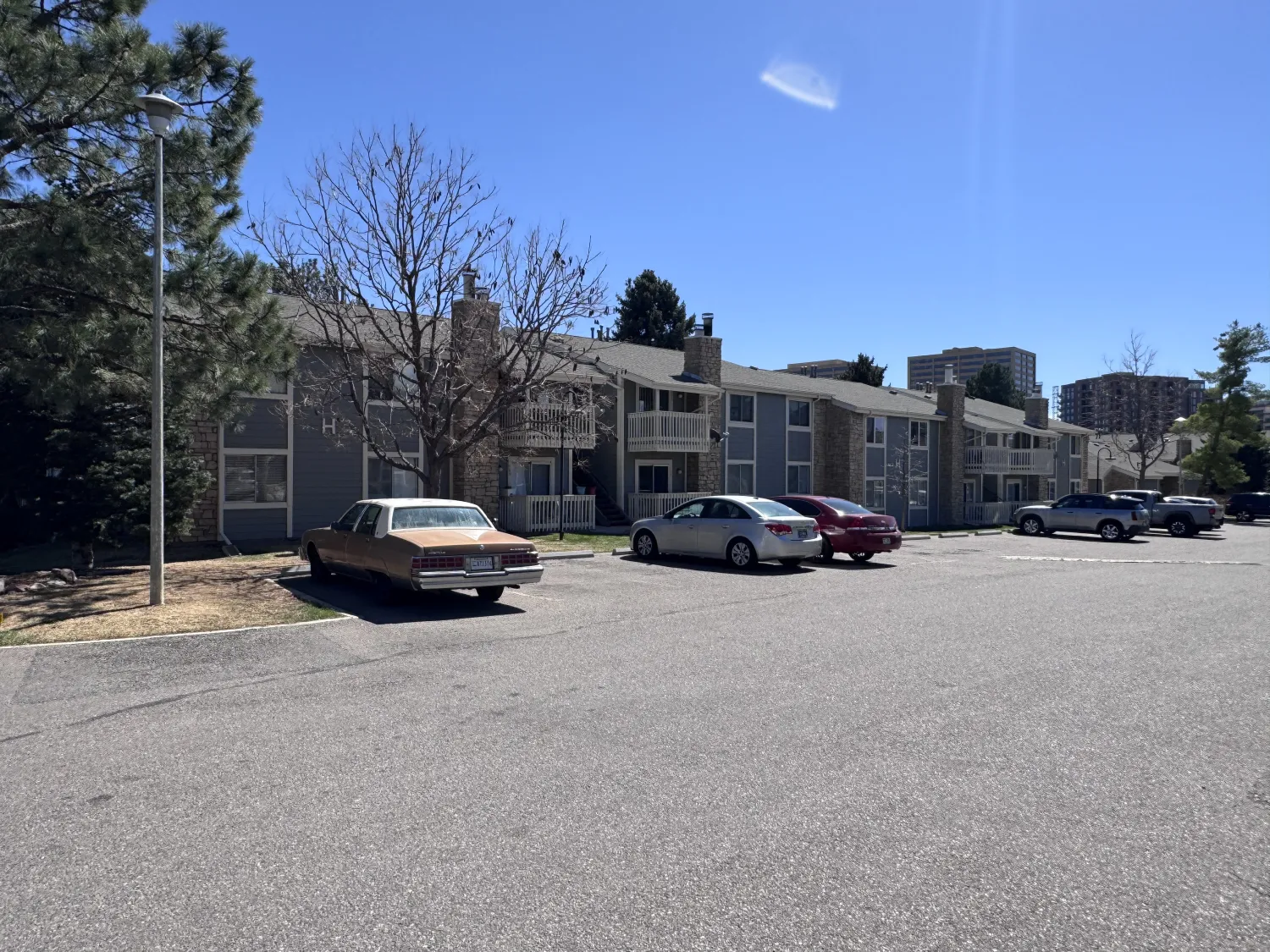 hoa-29 Apartment buildings with parked cars in front under a clear blue sky, trees, and a nearly empty parking lot.