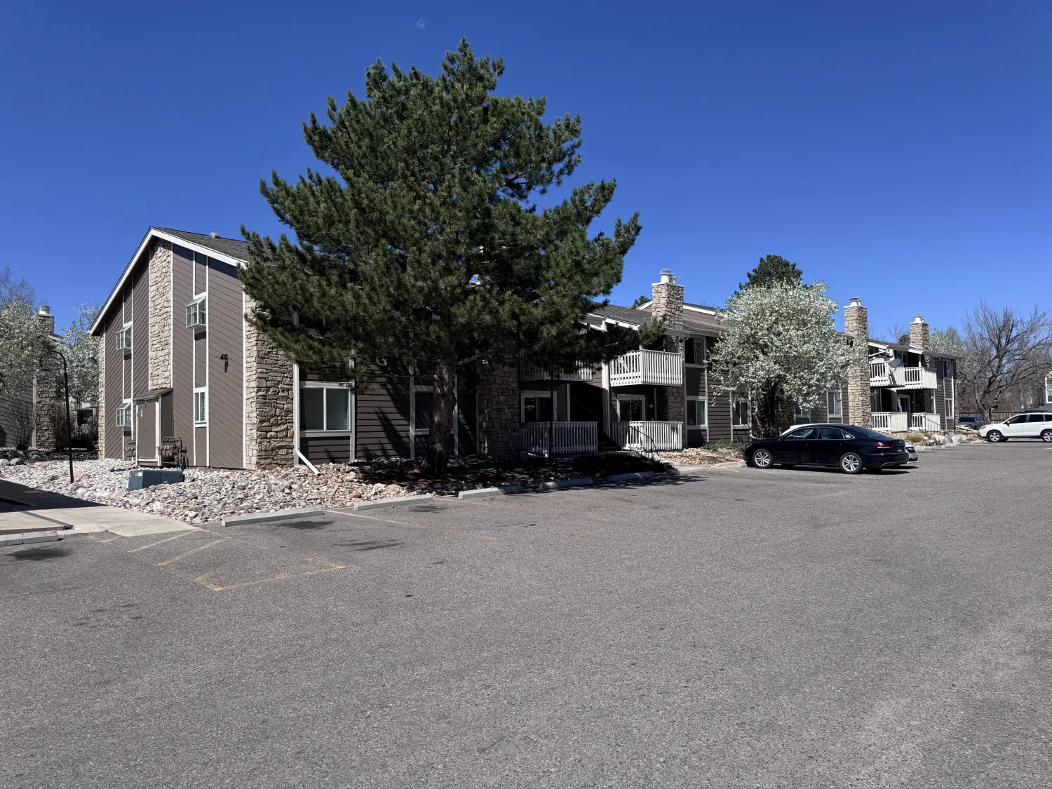 hoa-30 Two-story apartment building with balconies, a large pine tree, and a parked car under a clear blue sky.
