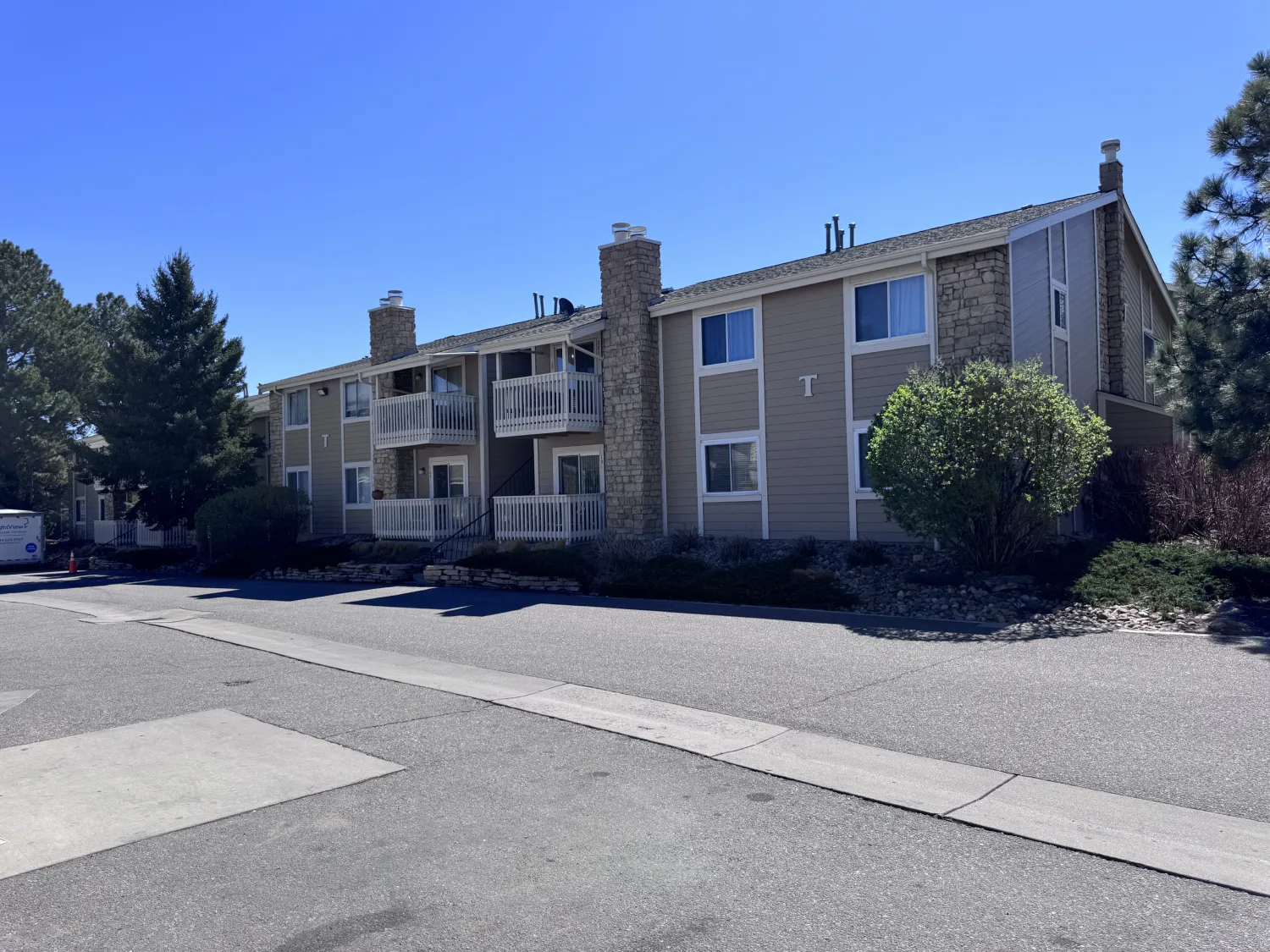 hoa-32 Two-story beige apartment building with balconies, stone chimneys, and trees along a quiet street.