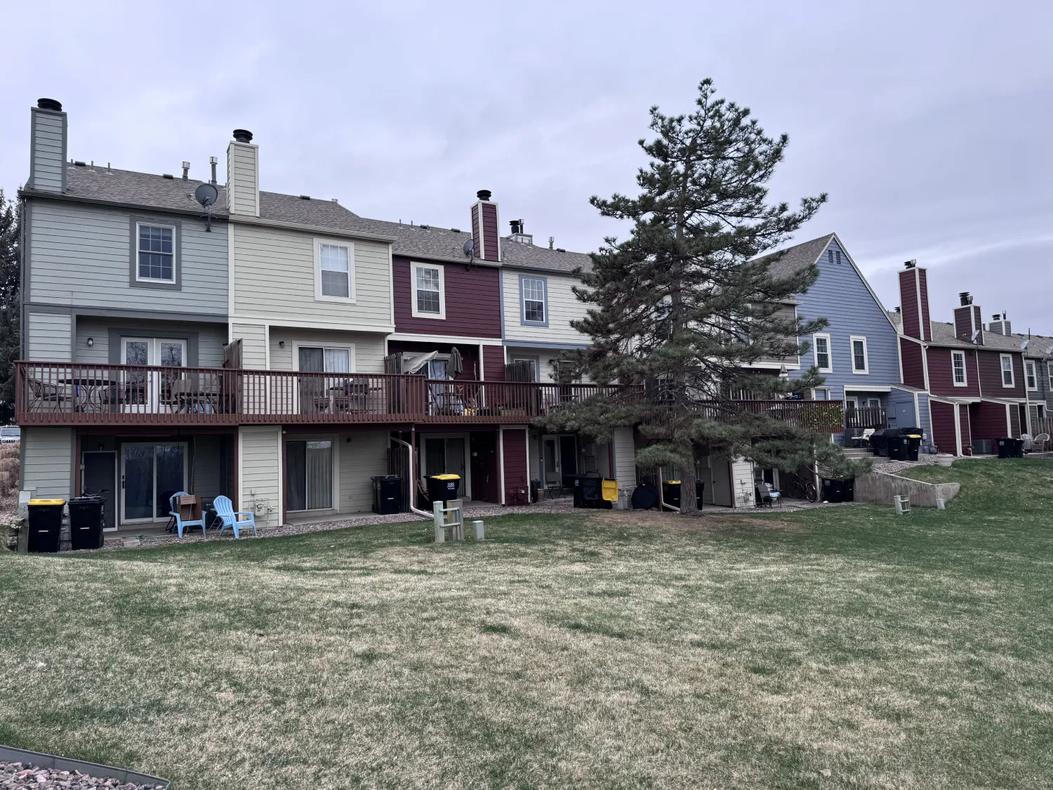 hoa-36 Row of multi-story townhomes with decks, lawn, patio furniture, and several trash bins on a cloudy day.