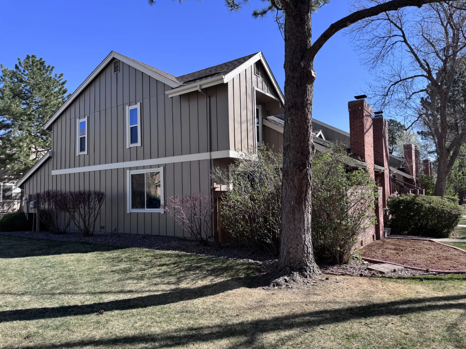 hoa-4 Gray two-story house with white trim, a brick chimney, trees, and bushes on a sunny day.