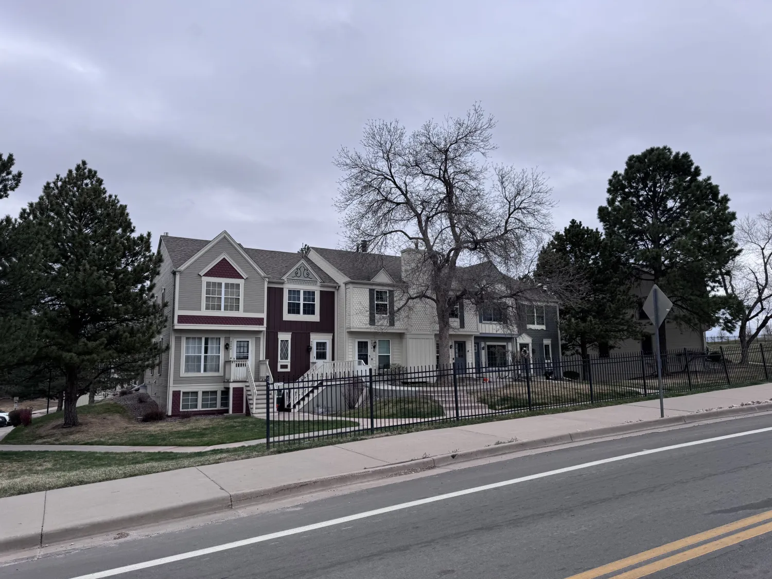 hoa-40 Townhouses with red and gray exteriors, trees, a fence, and a cloudy sky along a quiet street.