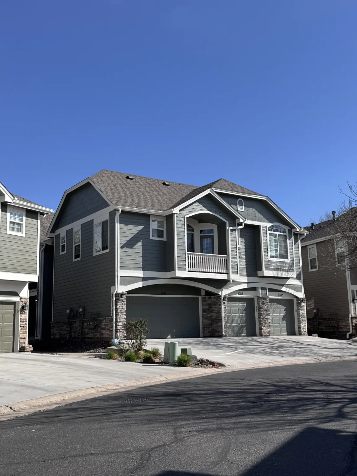 hoa-41 Two-story grey house with white trim, stone accents, balcony, three garages, and clear blue sky.