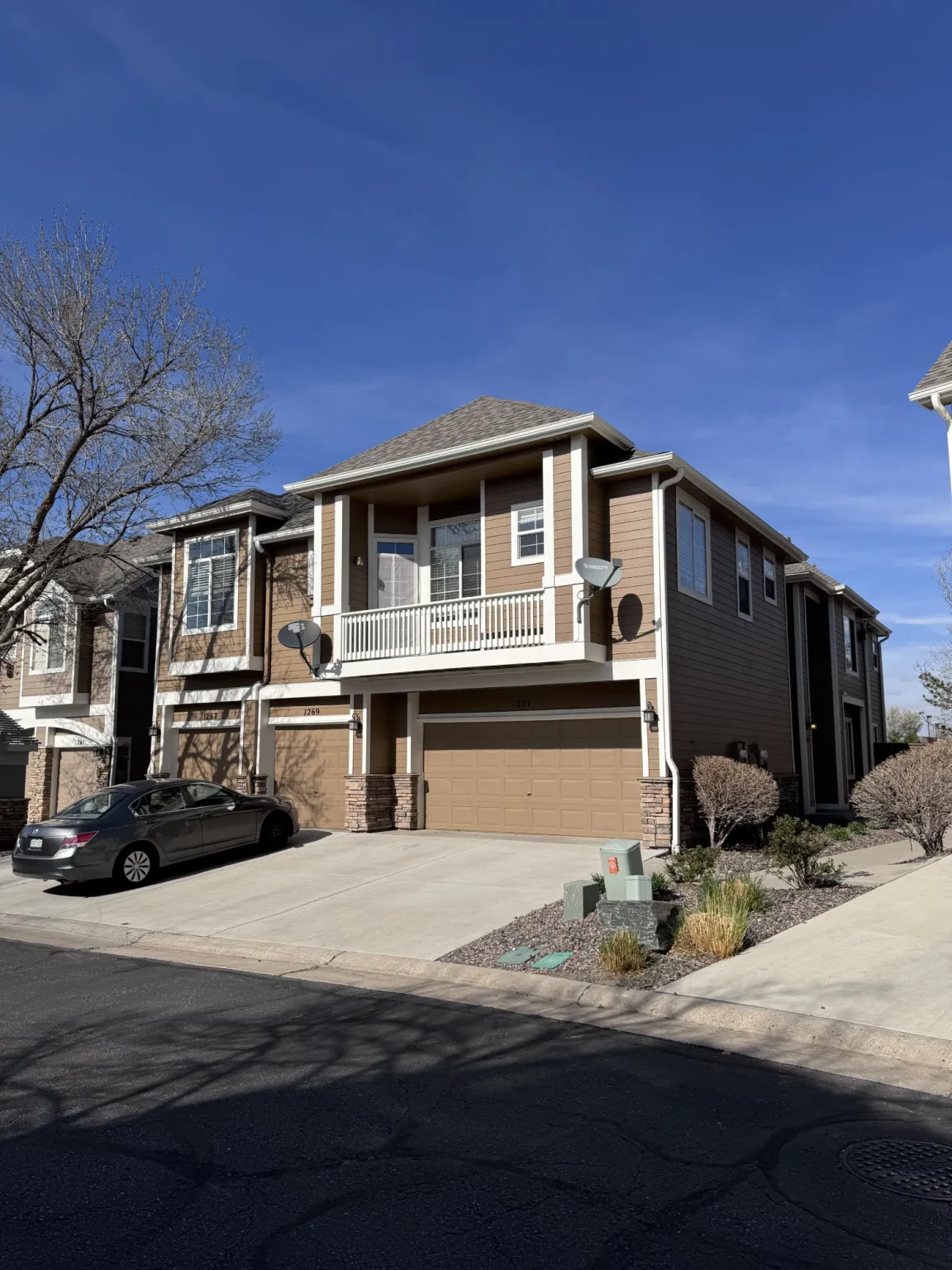 hoa-44 Two-story brown house with a balcony, double garage, parked car, and clear blue sky.