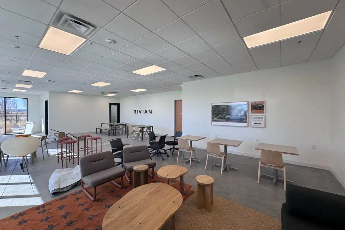 Modern office lounge with chairs, tables, and "RIVIAN" on the wall; large windows on the left.