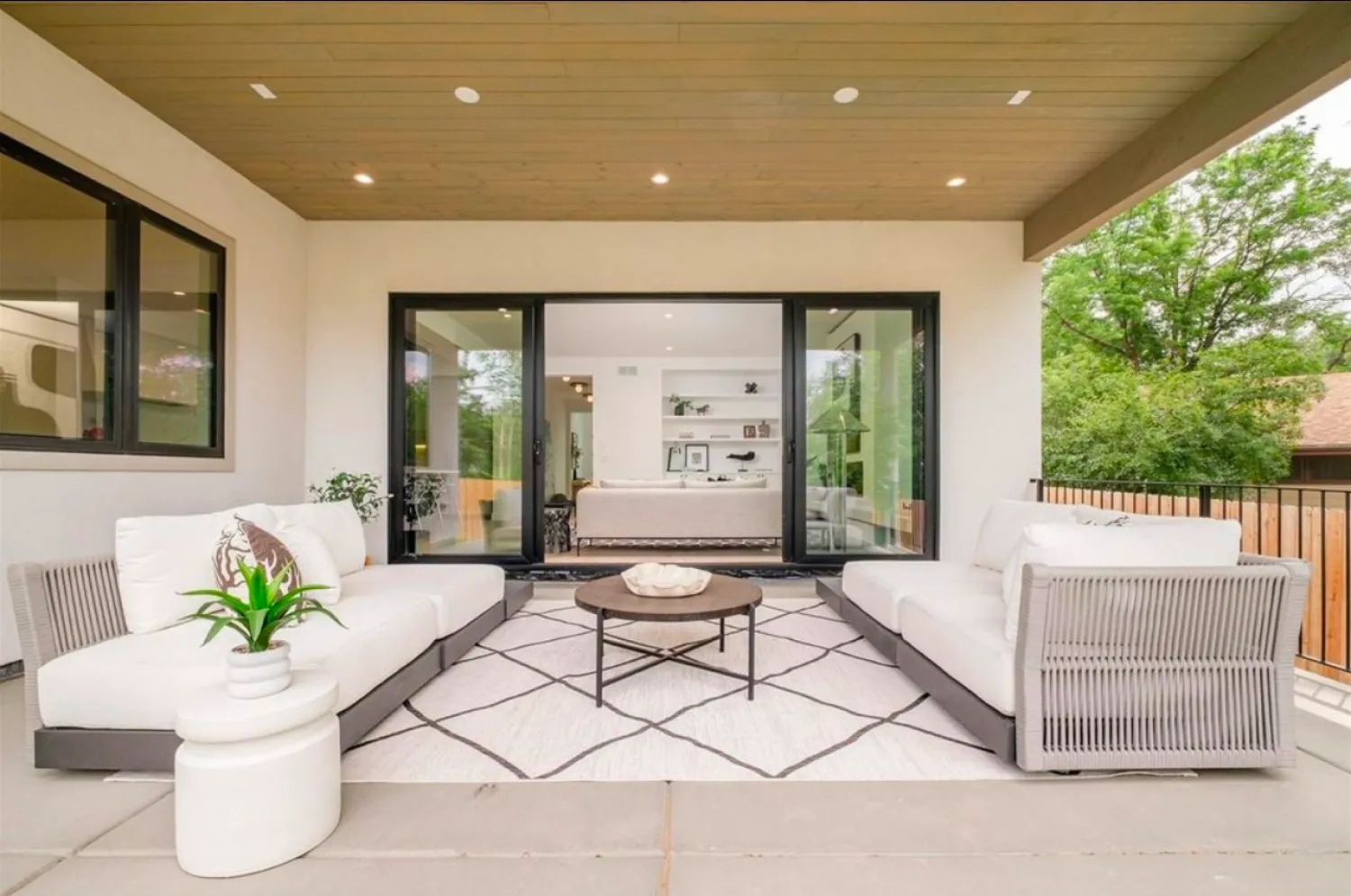 residential-exterior-16 Modern patio with white sofas, a round table, and a rug, overlooking a bright, open indoor living space.