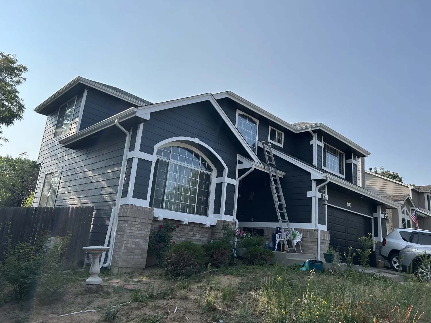 residential-exterior-20 Two-story dark blue house with white trim, a large front window, ladder, and overgrown yard.
