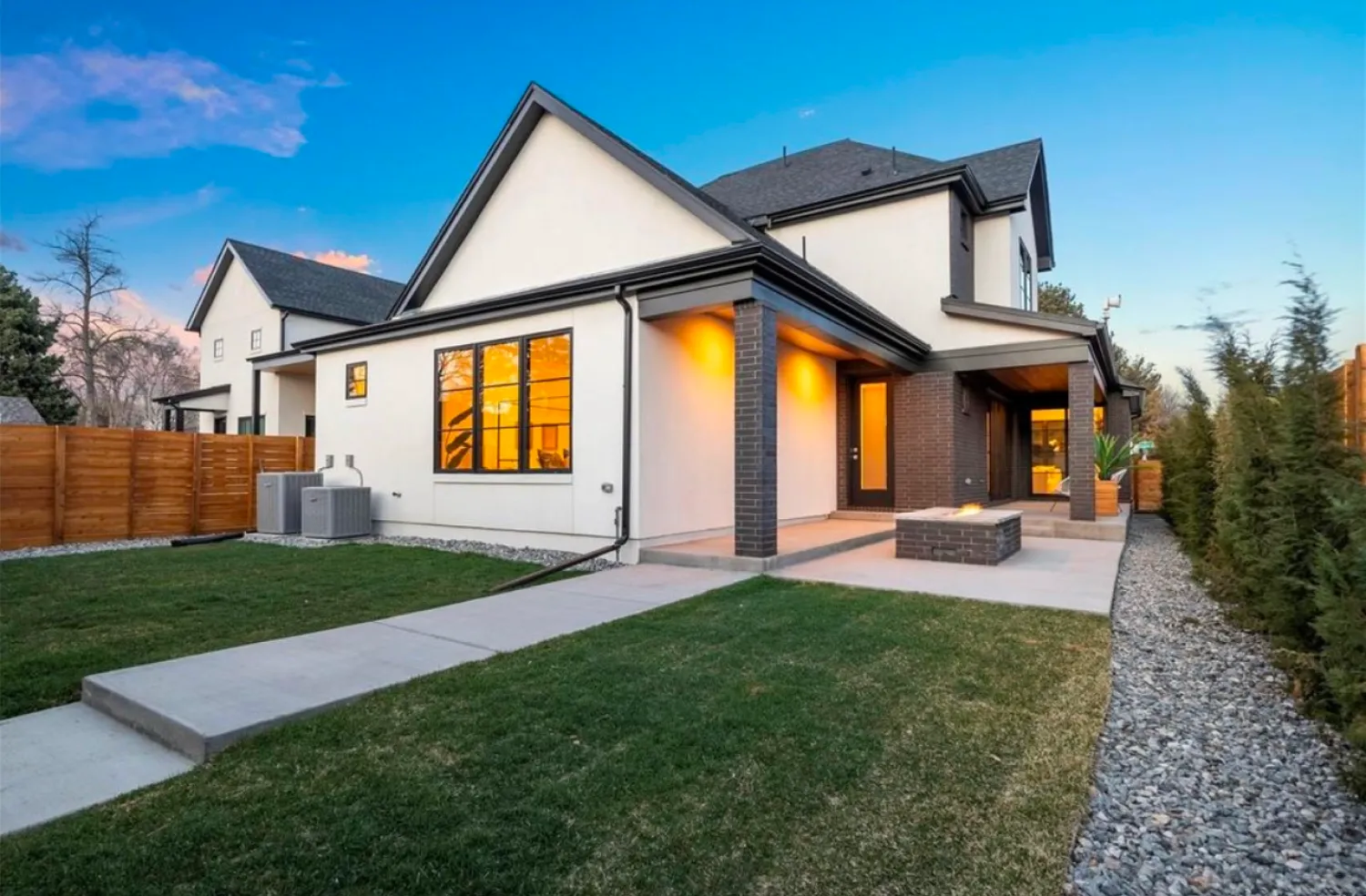 residential-exterior-3 Modern two-story house with large windows, porch lights on, and a fenced backyard at sunset.