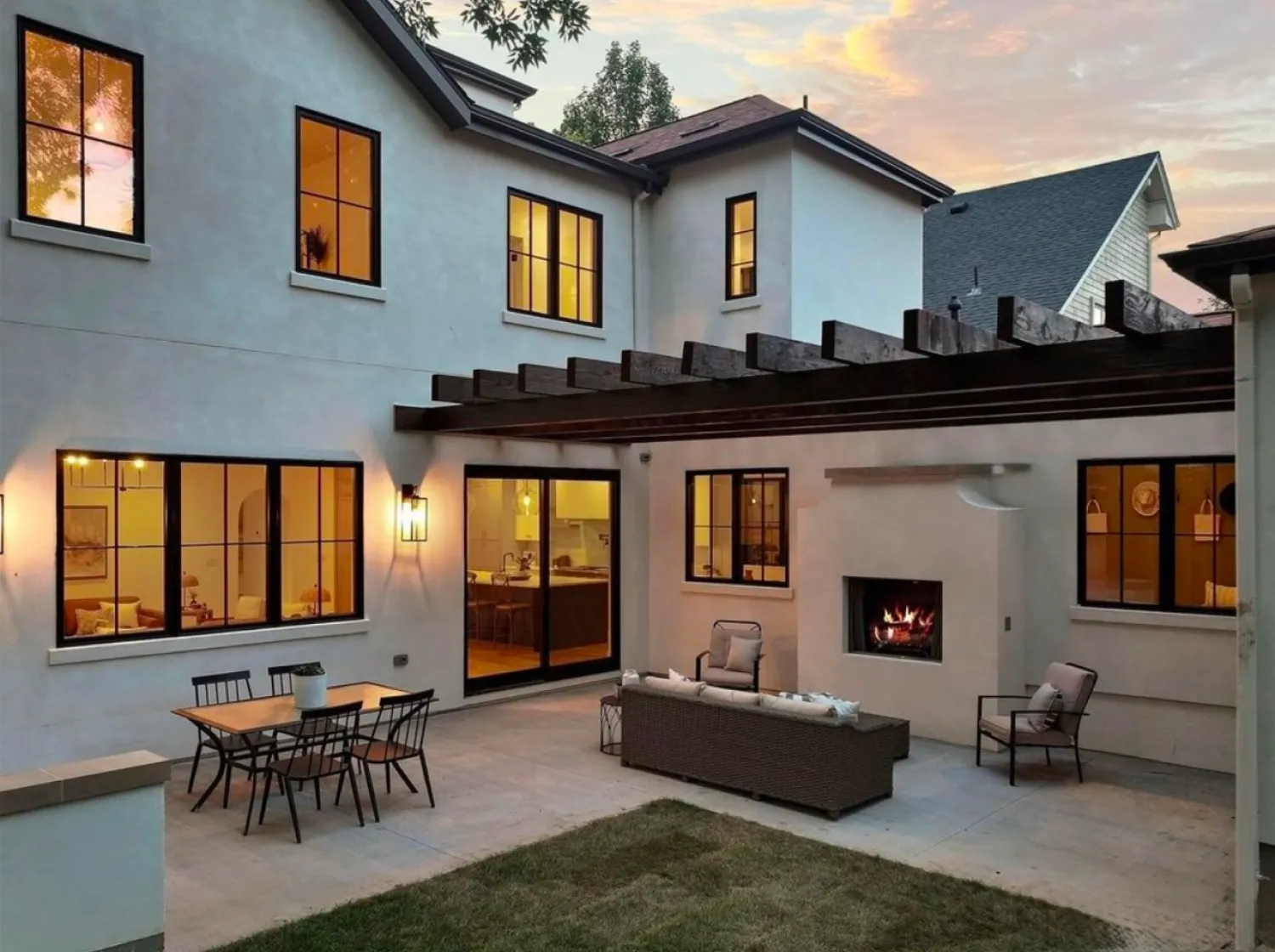 residential-exterior-6 Modern patio with outdoor sofa, dining table, fireplace, and pergola at sunset; house windows lit warmly.