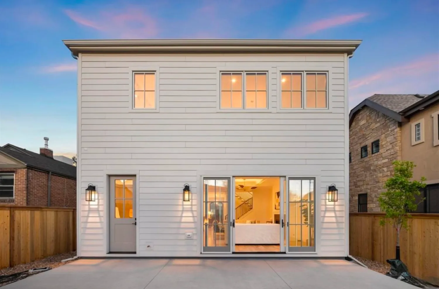 residential-exterior-8 Modern two-story white house with large windows, glass doors, and a fenced backyard at sunset.