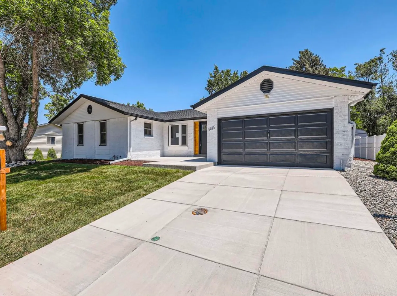 residential-exterior-9 White single-story house with black garage door, a green lawn, and a tree under a clear blue sky.