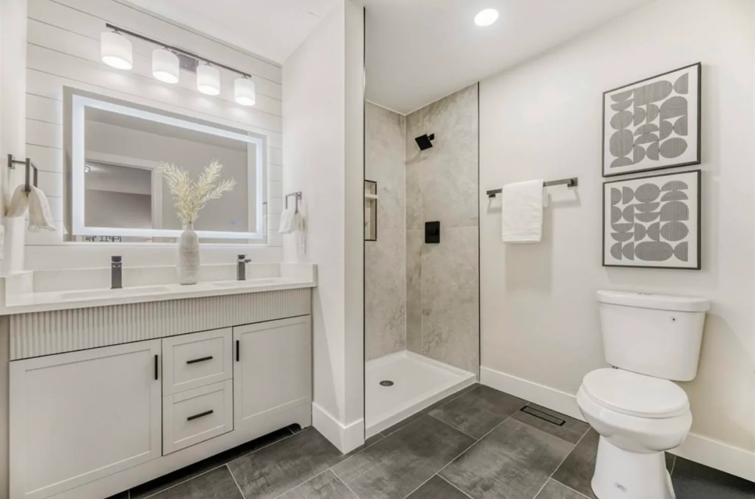 residential-interior-19 Modern bathroom with a double sink, glass shower, toilet, and two abstract art pieces on the wall.