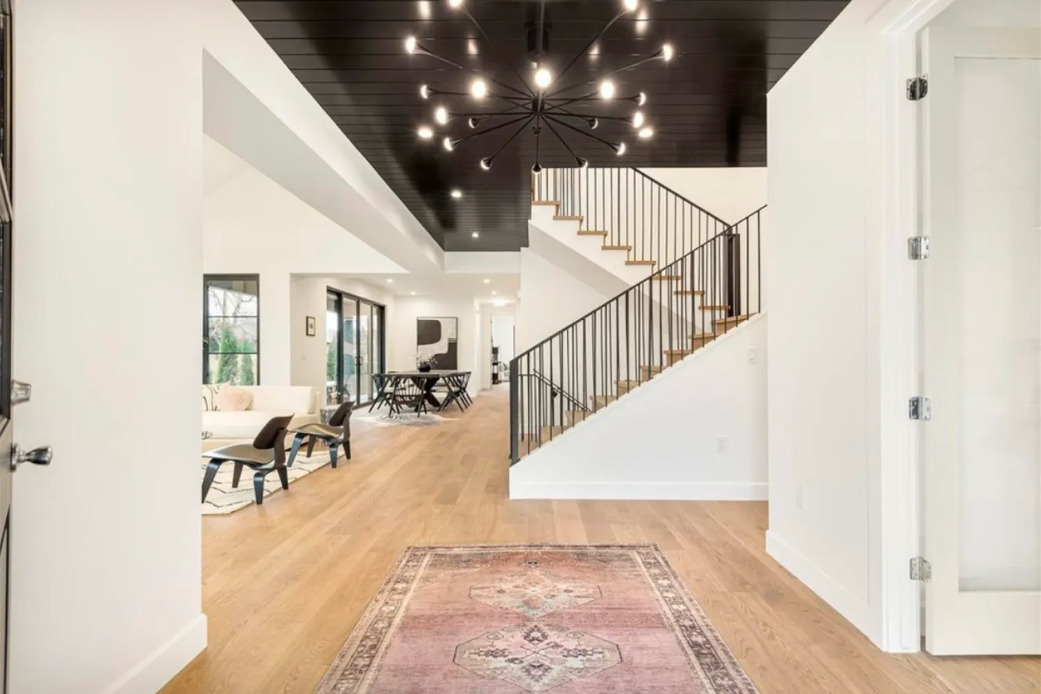 residential-interior-2 Modern open living space with wood floors, a staircase, black chandelier, and a patterned rug in the entryway.