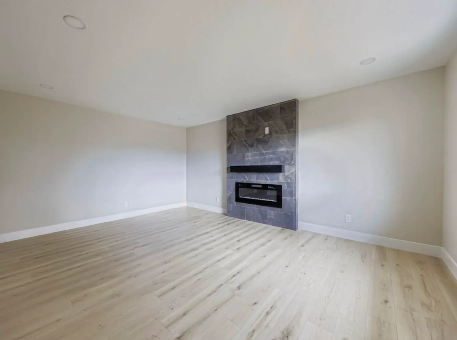 residential-interior-26 Empty room with light wood floor, beige walls, and a wall-mounted electric fireplace with a gray tile surround.