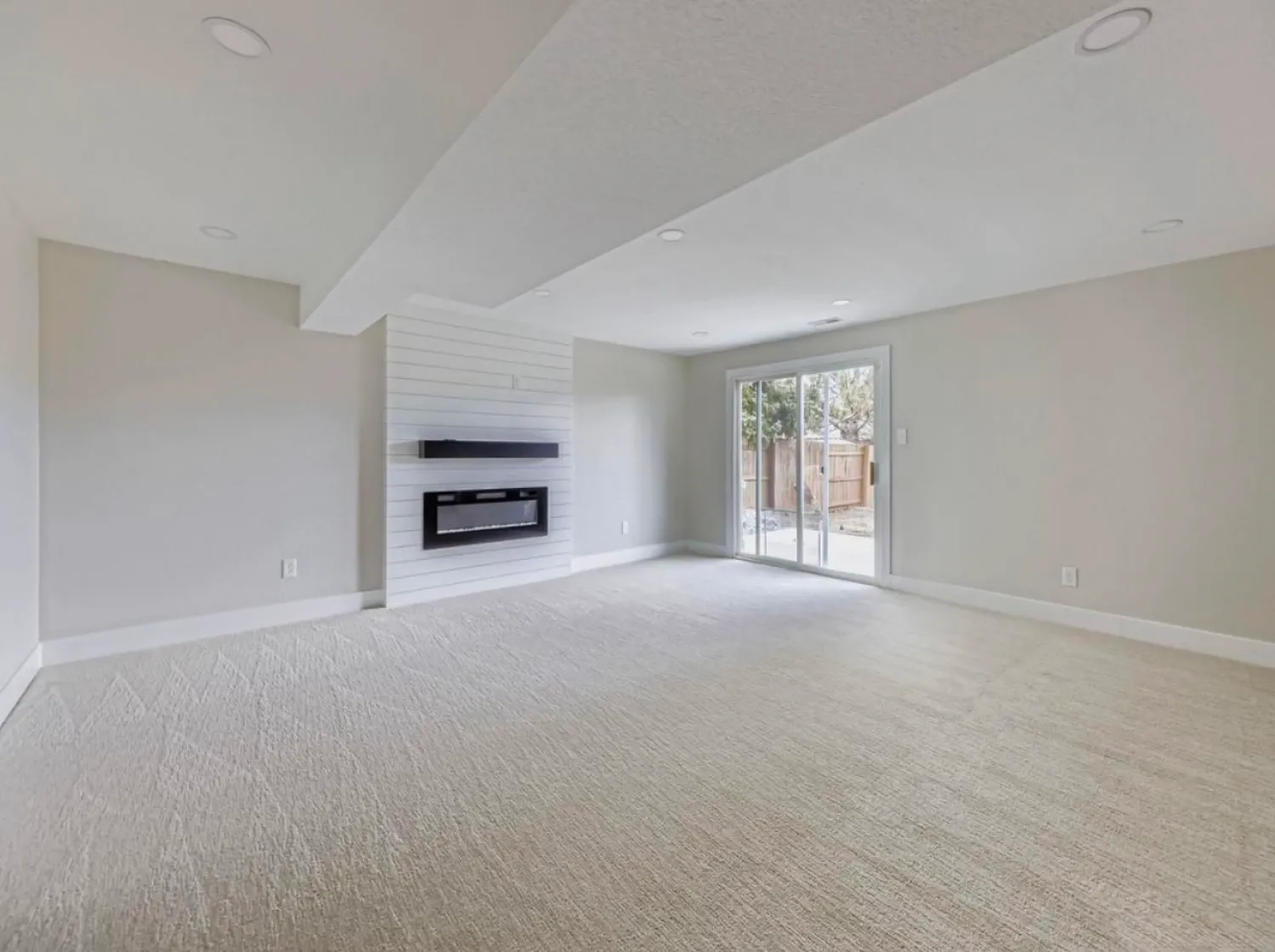 residential-interior-27 Spacious empty room with light carpet, wall-mounted fireplace, and glass door leading to a fenced backyard.