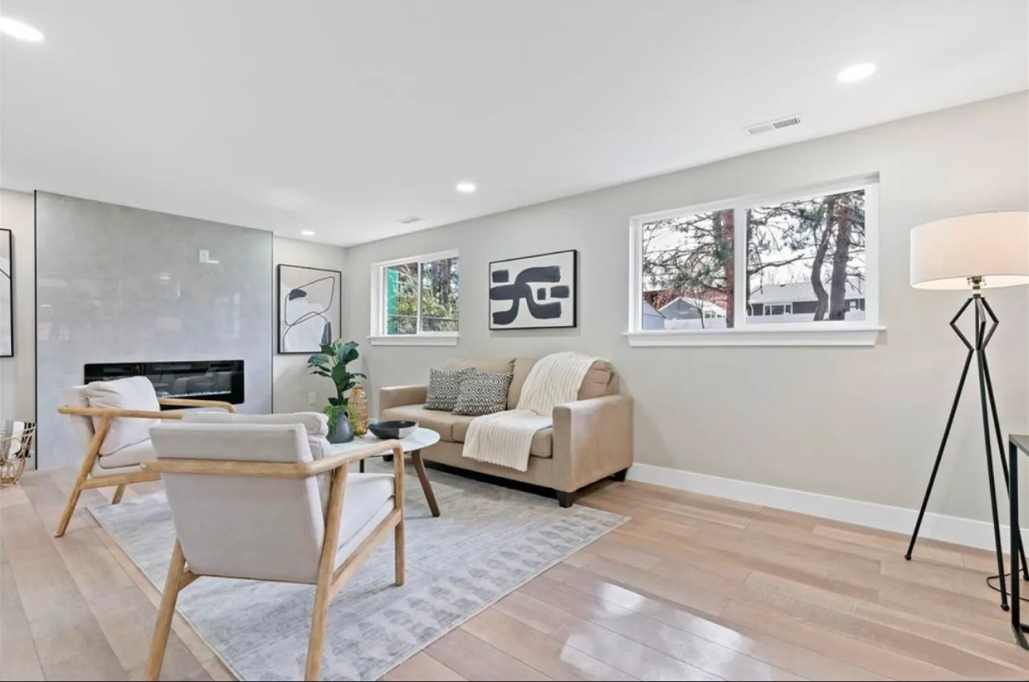 residential-interior-30 Modern living room with light wood floors, beige sofa, two chairs, wall art, and large windows.
