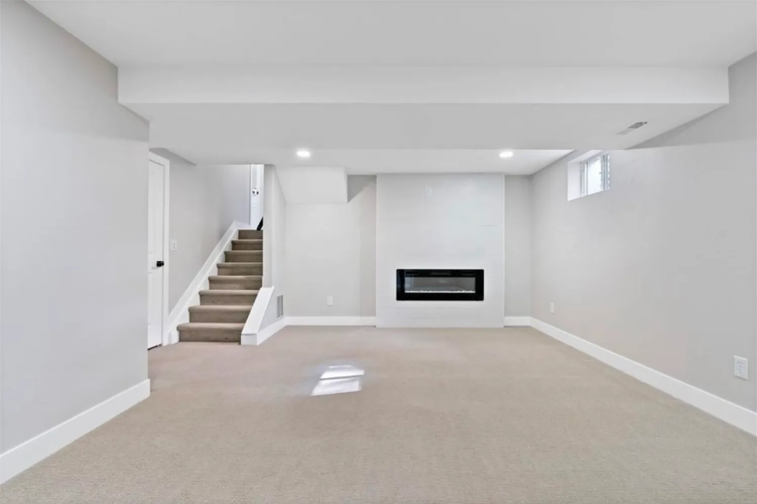 residential-interior-31 Modern basement with beige carpet, white walls, fireplace, small window, and stairs leading up.