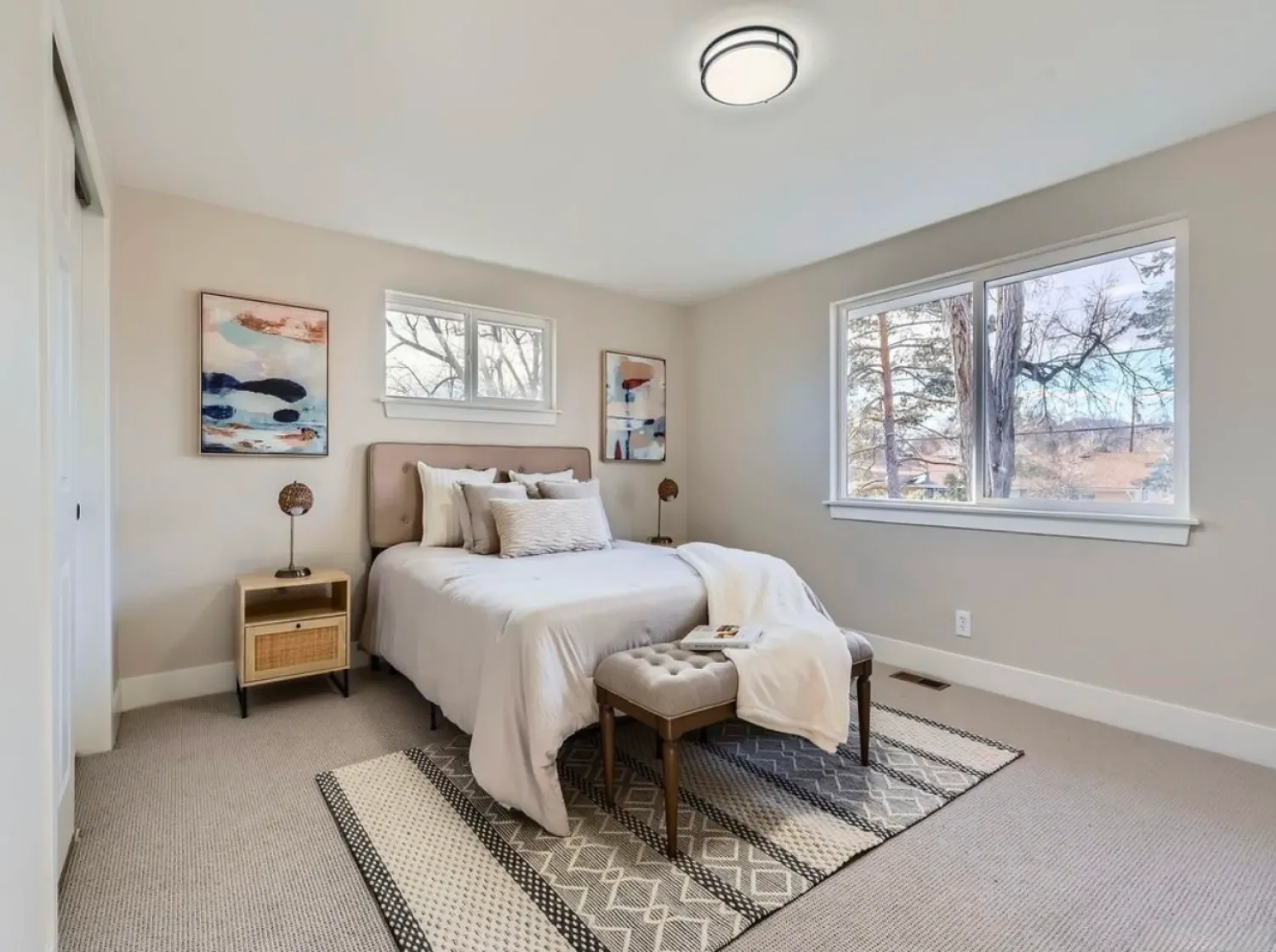 residential-interior-33 Bright bedroom with a double bed, two nightstands, a bench, and large windows showing trees outside.