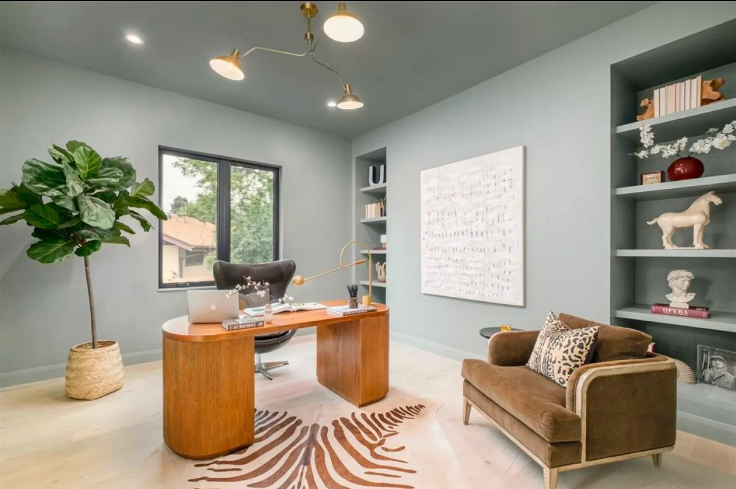 residential-interior-35 Modern home office with a wood desk, brown chair, zebra rug, shelves, and a large potted plant by the window.