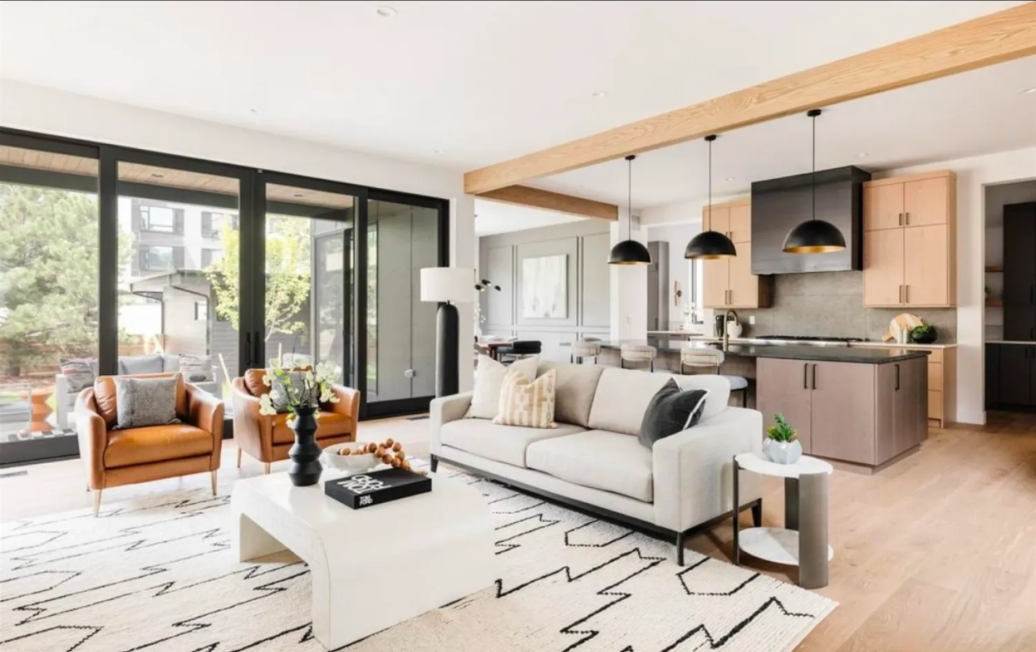 residential-interior-43 Modern living room with large windows, beige sofa, armchairs, and open kitchen with black pendant lights.