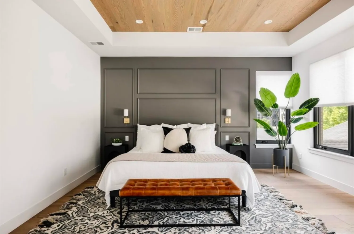residential-interior-44 Modern bedroom with a dark accent wall, white bedding, a brown bench, large plant, and wood ceiling.