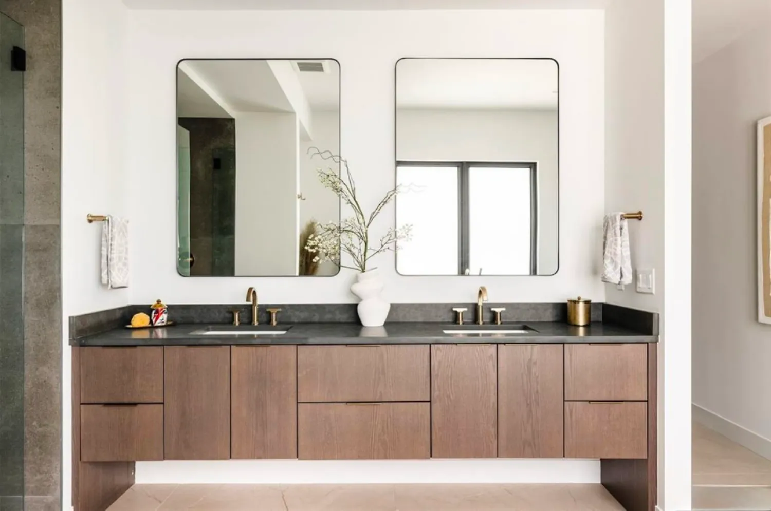 residential-interior-46 Modern bathroom with double sinks, wood vanity, two mirrors, brass fixtures, and a white vase with branches.
