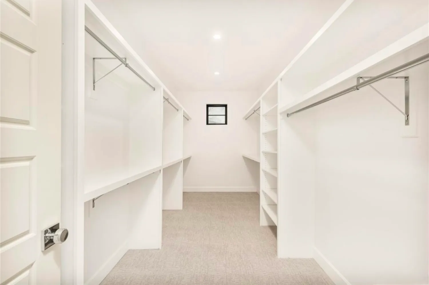 residential-interior-5 Spacious empty walk-in closet with white shelves, hanging rods, beige carpet, and a small window.
