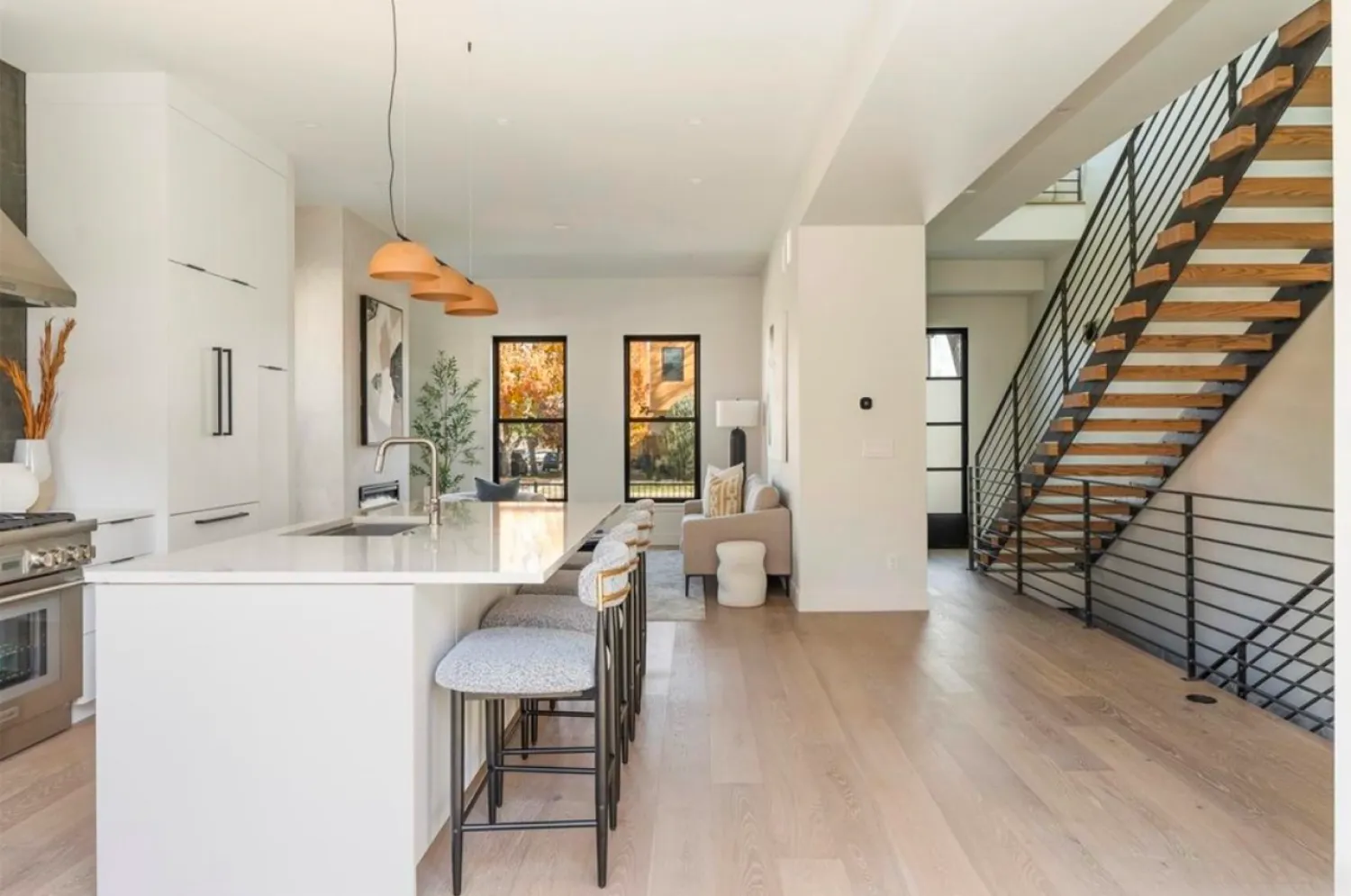 residential-interior-53 Modern kitchen with island seating, open living area, large windows, light wood floor, and metal staircase.