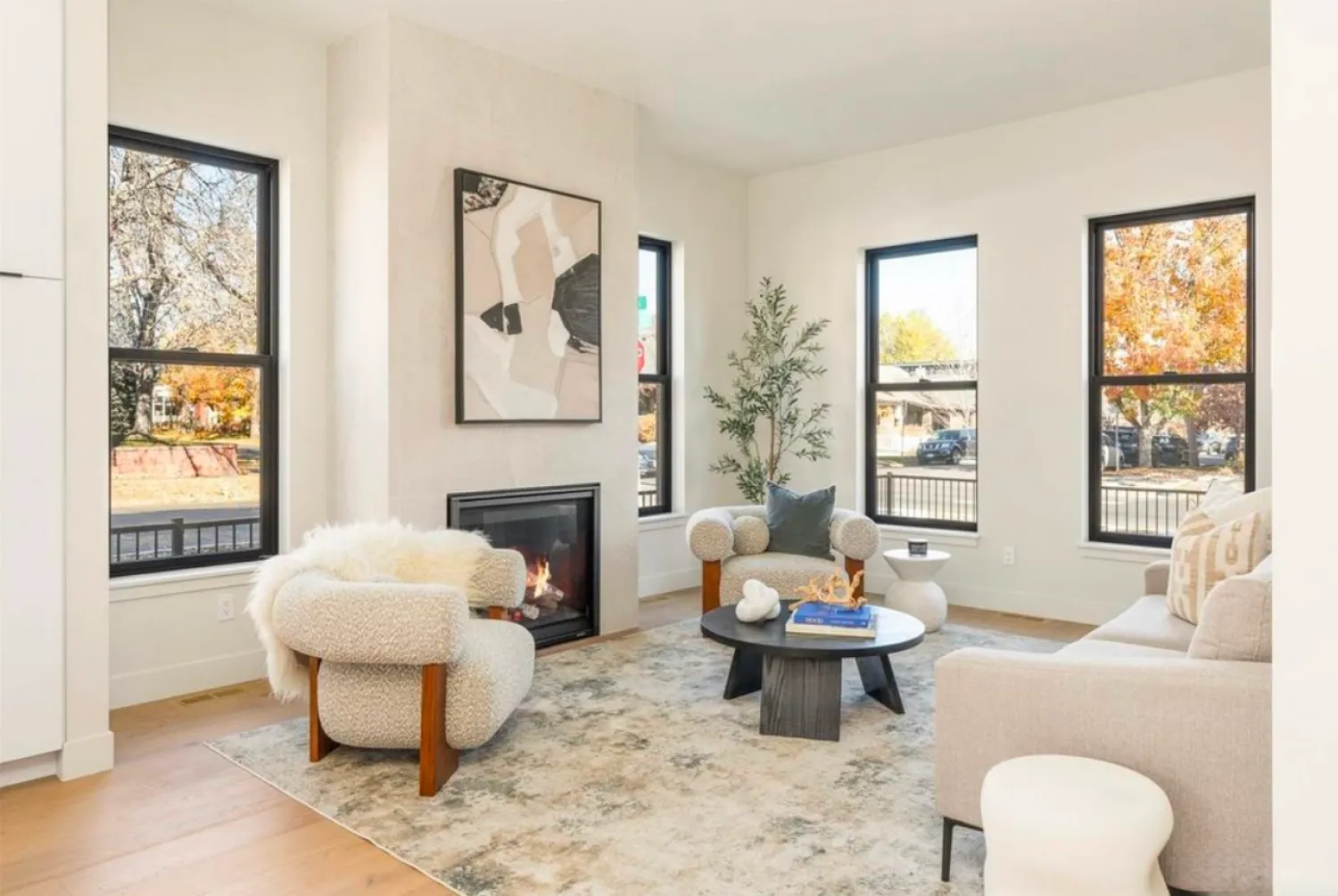 residential-interior-56 Bright living room with modern furniture, a fireplace, wall art, large windows, and autumn trees outside.