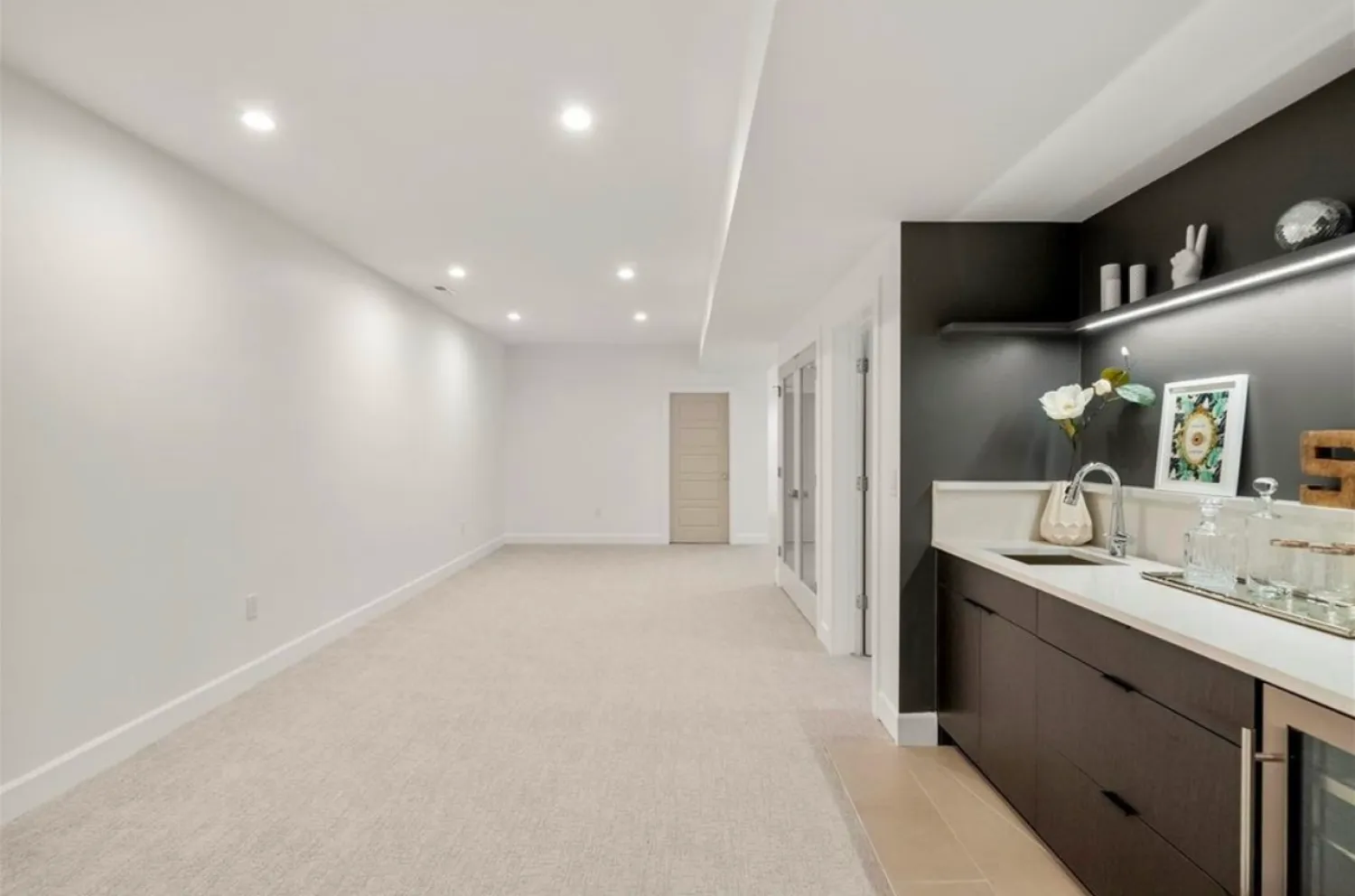 residential-interior-6 Modern, spacious basement with recessed lighting, beige carpet, and a sleek wet bar area with shelves and decor.