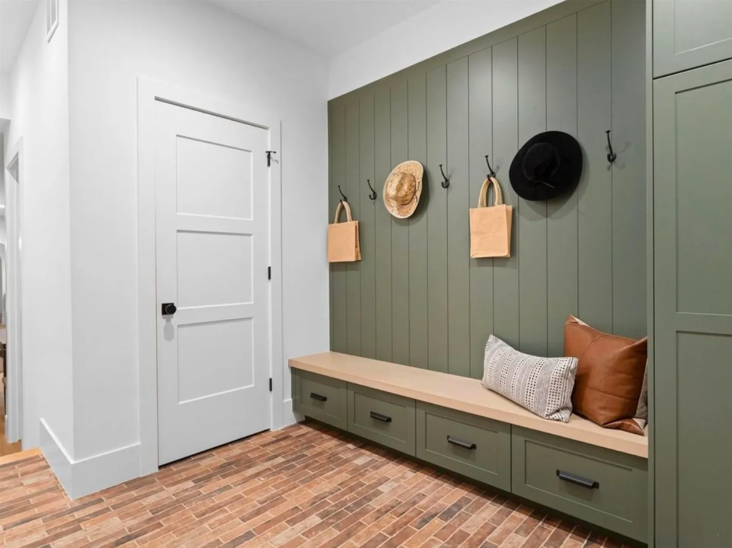 residential-interior-7 Mudroom with green built-ins, bench, hooks holding hats and bags, brick floor, and accent pillows.