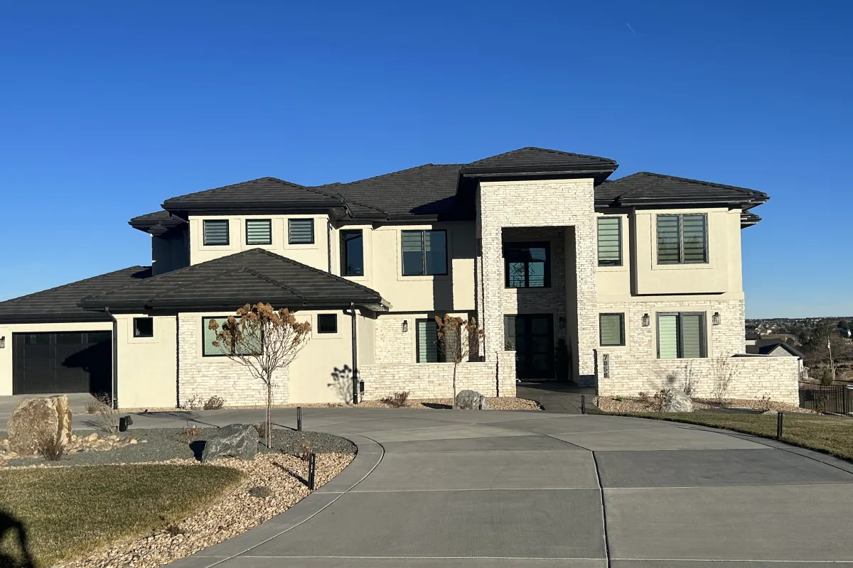 Modern two-story house with light stone exterior, black roof, and a curved driveway under a clear blue sky.